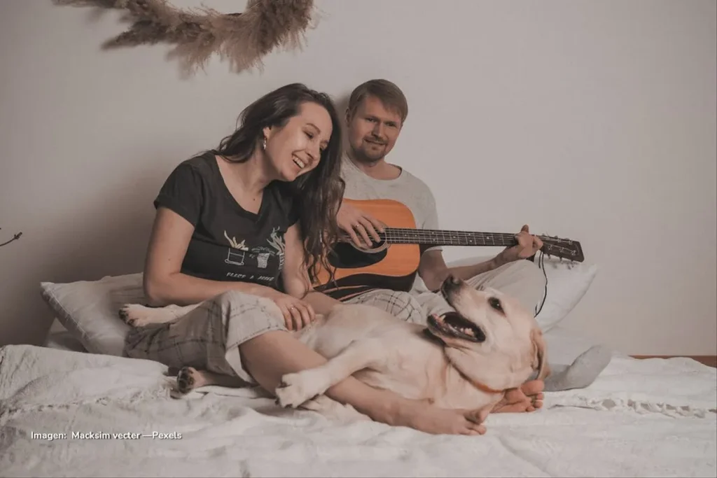 Mujer riendo junto a un perro recostado mientras un hombre toca la guitarra sobre la cama.